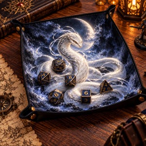 May include: A square black leather dice tray with gold-tone metal corner snaps, featuring a printed graphic of a white serpent coiled under a full moon, containing five dark blue polyhedral dice with gold numbers.