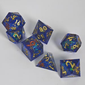 Puede incluir: Un juego de siete dados poliédricos de resina azul oscuro con números dorados y motivos celestiales. Los dados presentan escamas iridiscentes incrustadas y varias formas geométricas, incluyendo un d20, d12, d10, d8, d6 y d4 con bordes definidos.