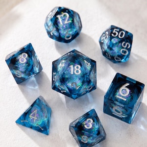 May include: A set of seven polyhedral dice made of translucent blue resin with embedded iridescent purple and green glitter flakes. Each die features sharp edges and clear, silver-coloured engraved numbers for high visibility during gameplay.