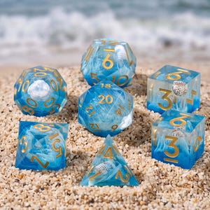 May include: A set of seven polyhedral dice made of clear blue resin with white inclusions resembling sea foam and small seashells embedded inside. Each die features gold-coloured numbers engraved on the faces, including a d20, d12, d10, d8, d6, d4, and a percentile die.
