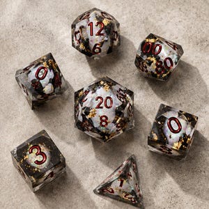 May include: A set of seven polyhedral dice made of clear resin with black inclusions, iridescent white flakes, and gold leaf accents. Each die features deep red engraved numbers, including a twenty-sided die, a twelve-sided die, and a percentile die.