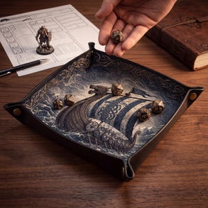 May include: A square leather dice tray featuring a printed illustration of a Viking longship sailing on turbulent blue waves. The tray has snap-button corners and contains several metallic polyhedral dice with gold-colored numbering.