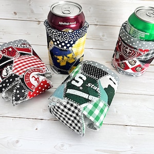 May include: Four fabric can coolers with patchwork designs. One cooler features the University of Alabama logo, another has the Michigan State logo, and a third has the University of Michigan logo. The fourth cooler has a red and white pattern. Each cooler holds a can.