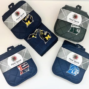 May include: A collection of quilted pot holders and a matching towel, each featuring a team logo. The pot holders and towel are navy blue, green, and red, with a plaid ribbon and the text "Plaid Spirit". The logos include "MICH", "Stats", and "DET LID".