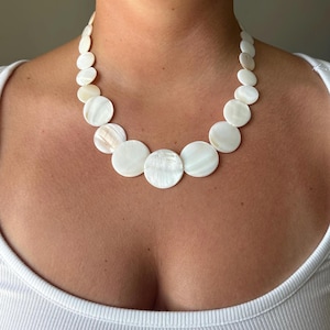 May include: A necklace composed of multiple circular, iridescent white shell discs. The discs are arranged in a graduated pattern, with the largest circular pieces positioned at the center and smaller discs extending toward the back of the neck.