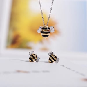Op de afbeelding: Een gouden en zwarte bij-hangerketting en bijpassende oorbellen. De bij is versierd met zwarte strepen en heeft zilveren vleugels.