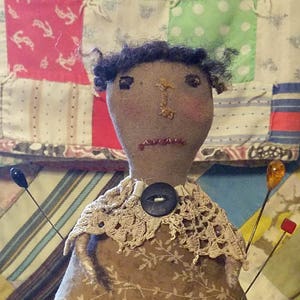 May include: A brown fabric pincushion doll with black hair, wearing a brown dress with white lace trim and a black button. The doll is sitting on a green box and has several pins sticking out of it.