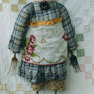 May include: A handmade rag doll with a plaid dress, a white apron with floral embroidery, and brown hair. The doll has fork legs and is standing on a white and brown patchwork quilt.
