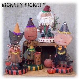 Może przedstawiać: Pięć ręcznie robionych dekoracji halloweenowych, w tym czarny kot, lalka z dynią na twarzy, czarownica i Święty Mikołaj. Dekoracje są wszystkie ubrane w świąteczne stroje i mają kapryśny, vintage'owy styl.