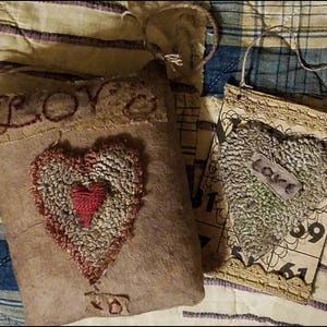 Op de afbeelding: Twee handgemaakte harten van stof met het woord "LOVE" erop geborduurd. Een hart is een kussen met een kleiner hart erin. Het andere hart is een wandhanger met een kantversiering.