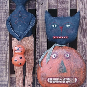 Op de afbeelding: Twee zwarte kattenpoppen van stof met gestikte details, waarvan er één een kleine oranje pompoen met zwarte sterren vasthoudt, worden getoond tegen een houten hek.