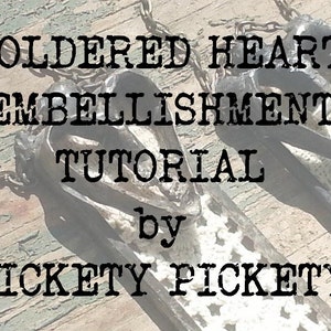 May include: A tutorial on how to make soldered heart embellishments, by Hickety Pickety. The image shows two black heart-shaped pendants with white lace details.