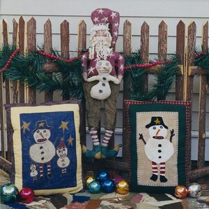 May include: Two handmade felt snowman pillows with a Santa Claus doll in the background. The pillows are decorated with stars, buttons, and a red and white striped pattern. The Santa Claus doll is wearing a red and white hat and a white beard.