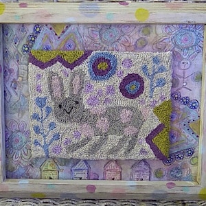 May include: A framed needlepoint artwork featuring a gray bunny with a pink and purple floral background. The frame is white with yellow and pink polka dots.