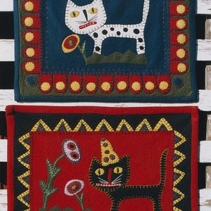 May include: Two framed needlepoint wall hangings featuring cats. The top one has a white cat with black spots on a blue background with a yellow flower. The bottom one has a black cat with a yellow hat on a red background with two red flowers.