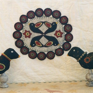 May include: Two black felt birds with red and yellow accents stand on wooden bases in front of a circular felt wall hanging with a design of four black birds with red and yellow accents.
