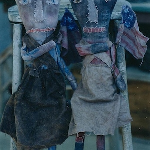 May include: Two rag dolls wearing crowns and holding American flags. The dolls are dressed in vintage-style clothing and have painted faces.
