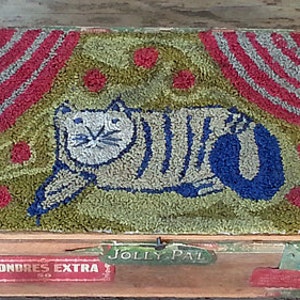 May include: A vintage wooden box with a colorful rug depicting a cat on a green background. The box has a label that reads "Londres Extra" and "Jolly Pal".