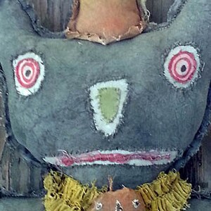 以下が含まれることがあります： グレーの顔、赤と白の目、緑の鼻をした手作りのかやの人形。人形は黄色のフリル襟を着ており、胸に小さなカボチャの顔が付いています。