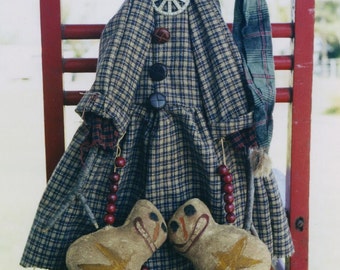 May include: A handmade rag doll wearing a plaid dress and a green and red plaid hat sits on a red wooden chair. The doll has a painted face with a wide smile and two small brown felt balls for eyes. The doll is holding two small brown felt figures with painted faces and a star shape on their bellies.