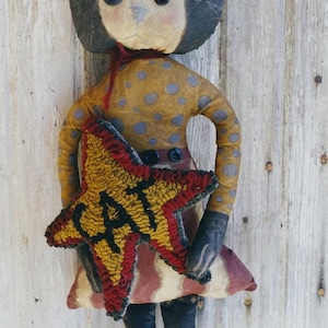 May include: A handmade rag doll cat with black ears and a brown and yellow star-shaped patch that says "CAT" on it. The doll is wearing a brown and white polka dot dress with red and white stripes on the bottom.