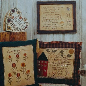 Op de afbeelding: Een hartvormige houten doos met de woorden "Mother's Treasures" erop geschilderd. De doos is versierd met bloemen en staat op een houten plank. Achter de doos hangt een ingelijst gedicht over een moeder aan de muur. Twee kussens met geborduurde ontwerpen liggen op een bed op de voorgrond. Het ene kussen heeft een huis en bloemen, het andere een gedicht over een moeder.