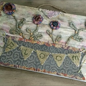 May include: A rustic wooden sign with a burlap background and a banner that reads "BLOOM". The sign is decorated with fabric flowers in shades of pink, purple, and brown.