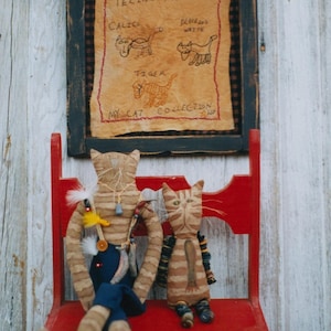 Könnte beinhalten: Zwei handgefertigte Katzenpuppen sitzen auf einem roten Holzstuhl. Die Puppen sind mit gestreiftem Stoff bekleidet und haben Knopfaugen. An der Wand hinter den Puppen hängt ein gerahmtes Stück Stoffkunst. Die Kunst zeigt ein handgesticktes Design von vier Katzen mit dem Text "Furry, Fuzzy, Funny Feline Friends" und "My Cat Collection".