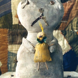 Pode incluir: Uma boneca de tecido cinza com uma boneca pássaro amarela presa ao seu peito. A boneca tem um nariz costurado e olhos de botão. A boneca pássaro tem uma cabeça amarela e um corpo verde.