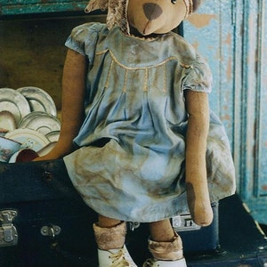 Peut inclure: Un ours en peluche brun portant une robe bleue avec des bordures en dentelle et des chaussures blanches est assis sur une valise vintage. L'ours a un nez brun et des yeux noirs.