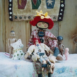 May include: A folk art display featuring a rag doll with a red hat and sunflower, a cat figurine, and small dolls. A wall hanging with mitten designs hangs in the background. The scene is set on a floral quilt.