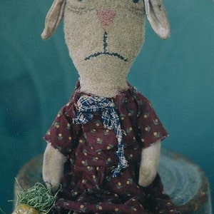 May include: A handmade rag doll with a brown face, plaid hat with a white flower, and a burgundy dress with a floral pattern. The doll is sitting on a weathered metal container.