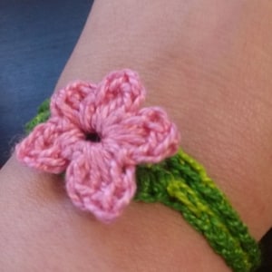 May include: A handmade crochet bracelet featuring a pink flower and green leaves. The flower is the focal point, with detailed petal stitching. The bracelet band is a vibrant green, creating a natural, floral theme.