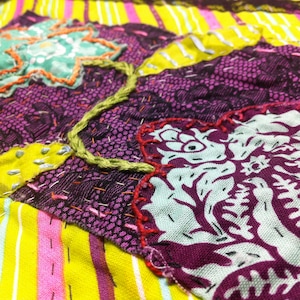 Puede incluir: Primer plano de un textil colorido con un diseño de patchwork. La tela presenta una mezcla de tonos morados, amarillos, rosas y verdes. Las costuras intrincadas y los detalles bordados añaden textura. El diseño incluye patrones florales y geométricos.