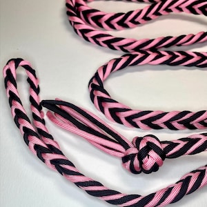 Może przedstawiać: Pleciona lina w kolorze różowym i czarnym. Lina jest ciasno tkana we wzór w jodełkę, z różowymi i czarnymi pasmami naprzemiennie, tworząc atrakcyjny wizualnie wzór. Lina jest zawiązana na jednym końcu, a drugi jest zapętlony.