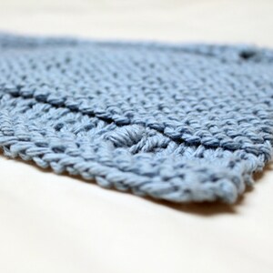 May include: Close-up of a light blue knitted item, possibly a scarf or blanket. The texture is visible, with a detailed stitch pattern. The item is lying on a white surface, with the focus on the knitted texture.