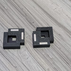 Peut inclure: Deux dessous de verre carrés noirs avec une surface texturée et des accents argentés. Les dessous de verre ont une forme emboîtable unique, créant une esthétique moderne et géométrique. Ils sont placés sur une surface en bois.