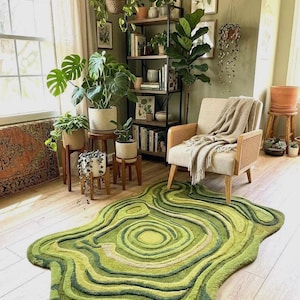 May include: A green rug with a topographic design in varying shades of green. The rug is placed on a light wood floor in a room with plants, a chair, and a bookshelf. The room has a natural, earthy aesthetic.