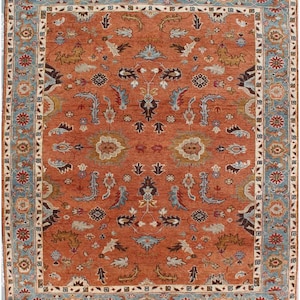 May include: A rectangular area rug featuring a burnt orange field with a symmetrical floral and vine pattern in shades of light blue, cream, and dark brown. The rug is framed by a wide border with repeating floral motifs on a light blue background.