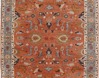 Könnte beinhalten: Ein rechteckiger Teppich mit einem rostorangen Grund und einem symmetrischen Blumen- und Rankenmuster in Hellblau, Creme und Dunkelbraun. Der Teppich ist von einer breiten Bordüre mit wiederkehrenden floralen Motiven auf hellblauem Grund umgeben.