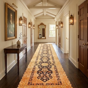 May include: A long, rectangular runner rug with a central black and cream floral pattern, bordered by a warm orange and gold design with fringed ends, positioned on a dark hardwood floor in a hallway with cream walls and wooden doors.
