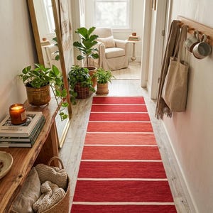 May include: A hallway runner rug with horizontal stripes in shades of red and white. The rug is placed in a hallway with a wooden bench, plants, and a mirror. The overall theme is home decor.