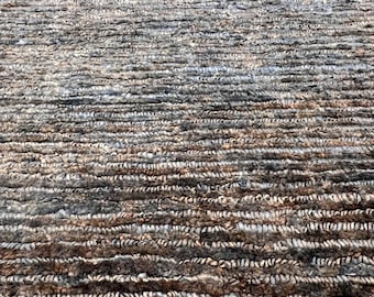 May include: Close-up of a textured rug with a pattern of horizontal lines. The rug features a blend of brown, gray, and beige tones, creating a natural, earthy aesthetic. The fibers appear to be tightly woven, suggesting durability.