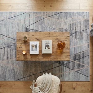 May include: Overhead view of a living room setup. A light-colored sofa with throw pillows sits next to a geometric patterned rug. A wooden coffee table holds books, a candle, and a decorative glass sphere. Potted plants are on the left.