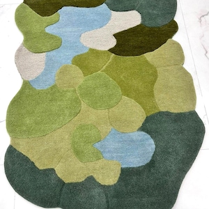 Overhead view of a hand-tufted wool rug with an irregular organic shape in shades of green and blue on a white marble floor.