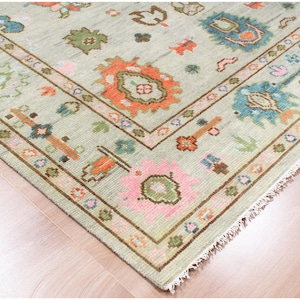 May include: A corner view of a light green rug with a detailed border. The rug features floral and geometric patterns in shades of orange, pink, blue, and brown. The rug is placed on a wooden floor.