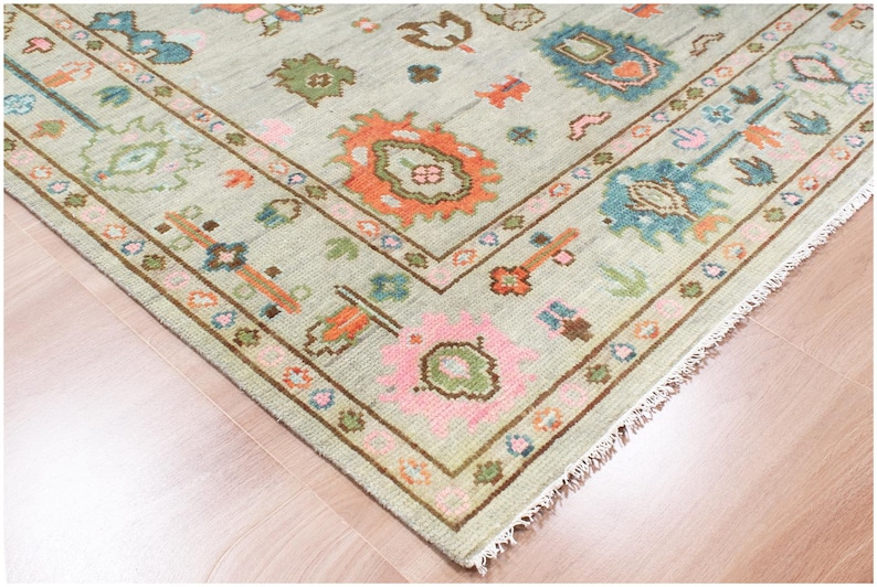 May include: A corner view of a light green rug with a detailed border. The rug features floral and geometric patterns in shades of orange, pink, blue, and brown. The rug is placed on a wooden floor.