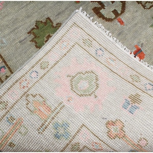 May include: Close-up of a section of a vintage rug with a light gray background. The rug features a detailed pattern with floral and geometric designs in shades of pink, blue, green, and brown. The edges are finished with a white fringe.