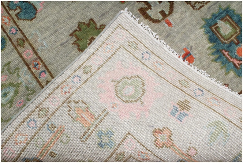 May include: Close-up of a section of a vintage rug with a light gray background. The rug features a detailed pattern with floral and geometric designs in shades of pink, blue, green, and brown. The edges are finished with a white fringe.