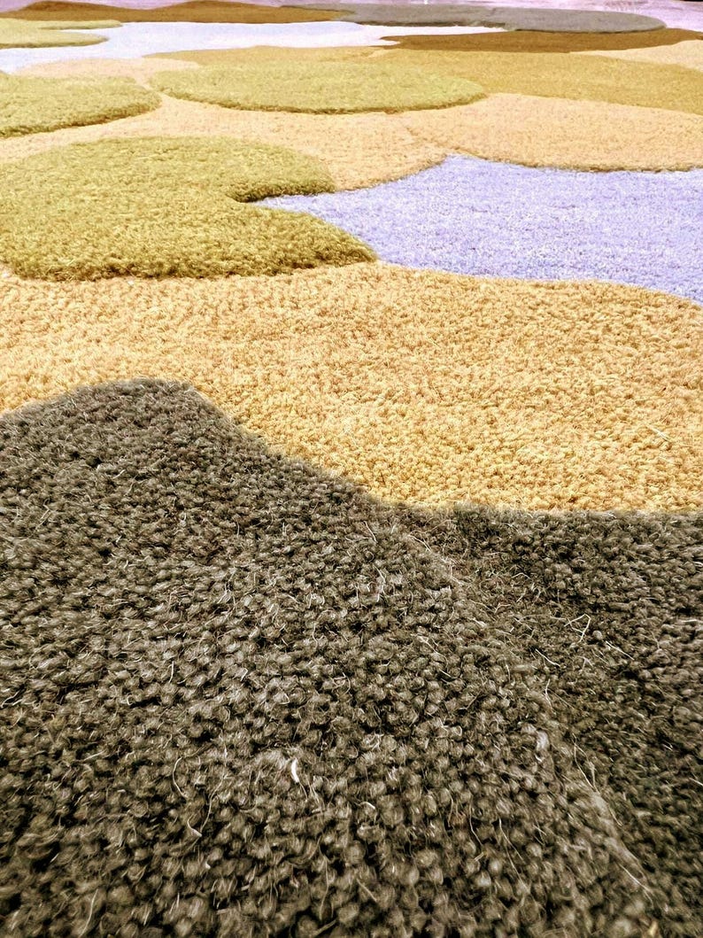 May include: A large, textured rug with a landscape design. The rug features various colors, including olive green, tan, and light purple, creating a soft, plush appearance. The design resembles a stylized landscape.